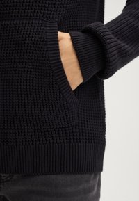 Ciemnoniebieski sweter o fakturowanym wzorze w splot wafelka, z boczną kieszonką oraz ściągaczami przy mankietach i na dolnym brzegu. Materiał wydaje się miękki i ciepły.