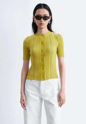 Kvinde iført sorte solbriller, gul ribbet kortærmet knappet top og hvide bukser, stående mod en lysegrå baggrund.