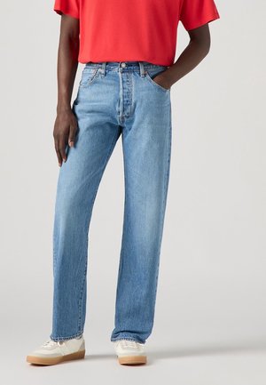 Personne portant un jean droit bleu clair, un t-shirt rouge à manches courtes, des baskets blanches, une main dans la poche et l'autre détendue le long du corps.