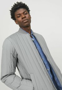 Giacca bomber trapuntata grigia con colletto a coste, chiusura con zip e tasche laterali, indossata sopra una camicia in denim blu.