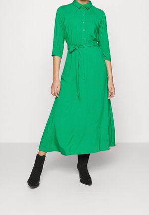 Vrouw die een lange groene jurk met knopen draagt, driekwartmouwen en een geknoopt tailleceintuur, gecombineerd met zwarte enkellaarzen.