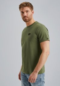 T-shirt in cotone verde oliva, design a girocollo, maniche corte, piccola etichetta nera sul petto, texture morbida, abbinato a jeans azzurri chiari.