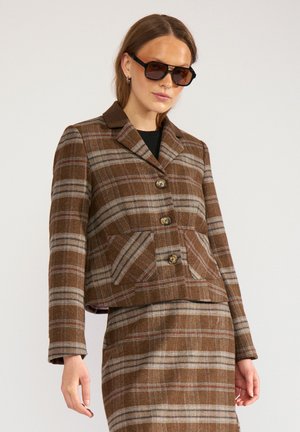 Femme portant une veste à carreaux marron et une jupe assortie avec des lunettes de soleil foncées sur un fond uni.