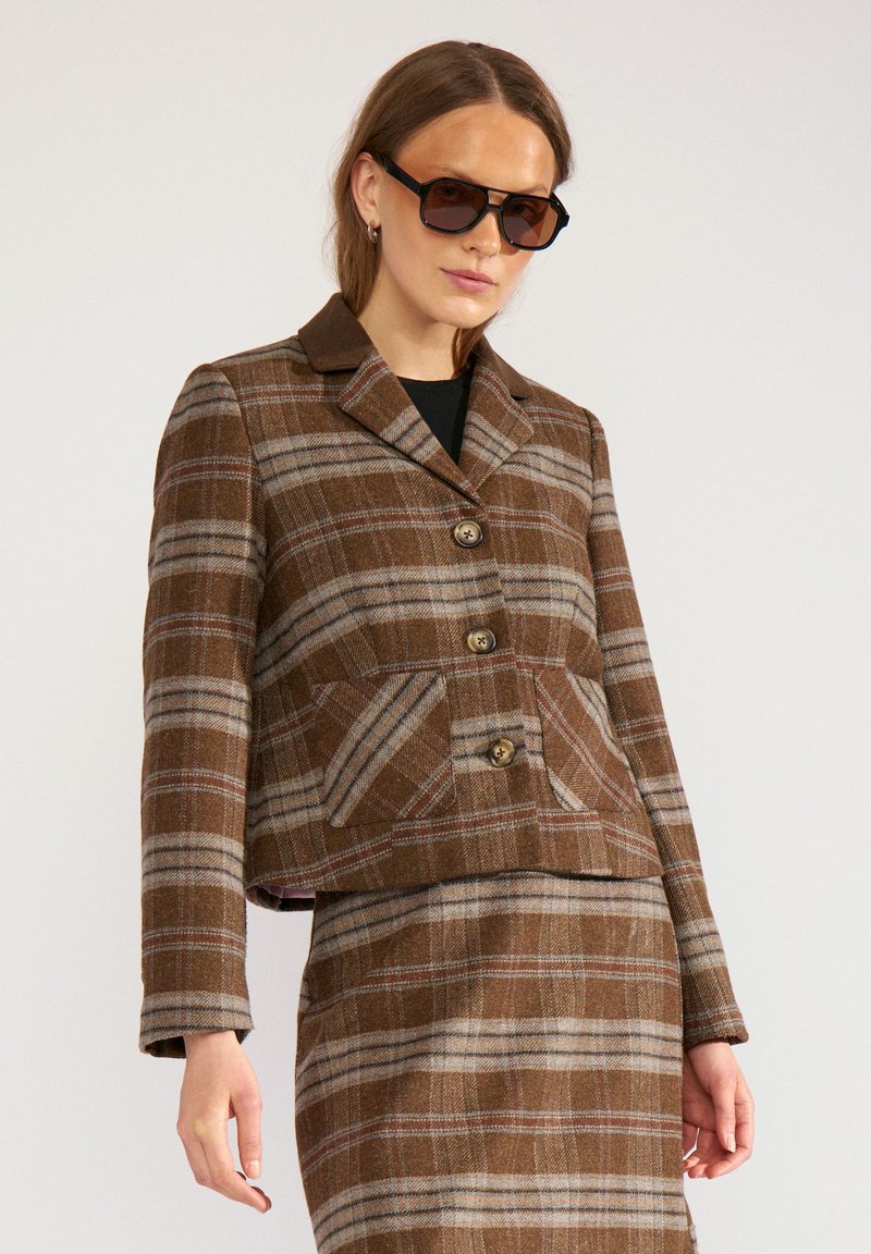 Femme portant une veste à carreaux marron et une jupe assortie avec des lunettes de soleil foncées sur un fond uni.