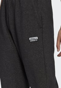 Tumši pelēki sporta bikses, kas izgatavotas no mīkstas auduma ar sānu kabatām. Uz kreisās puses ir melnbalta Adidas logo uzlīme.