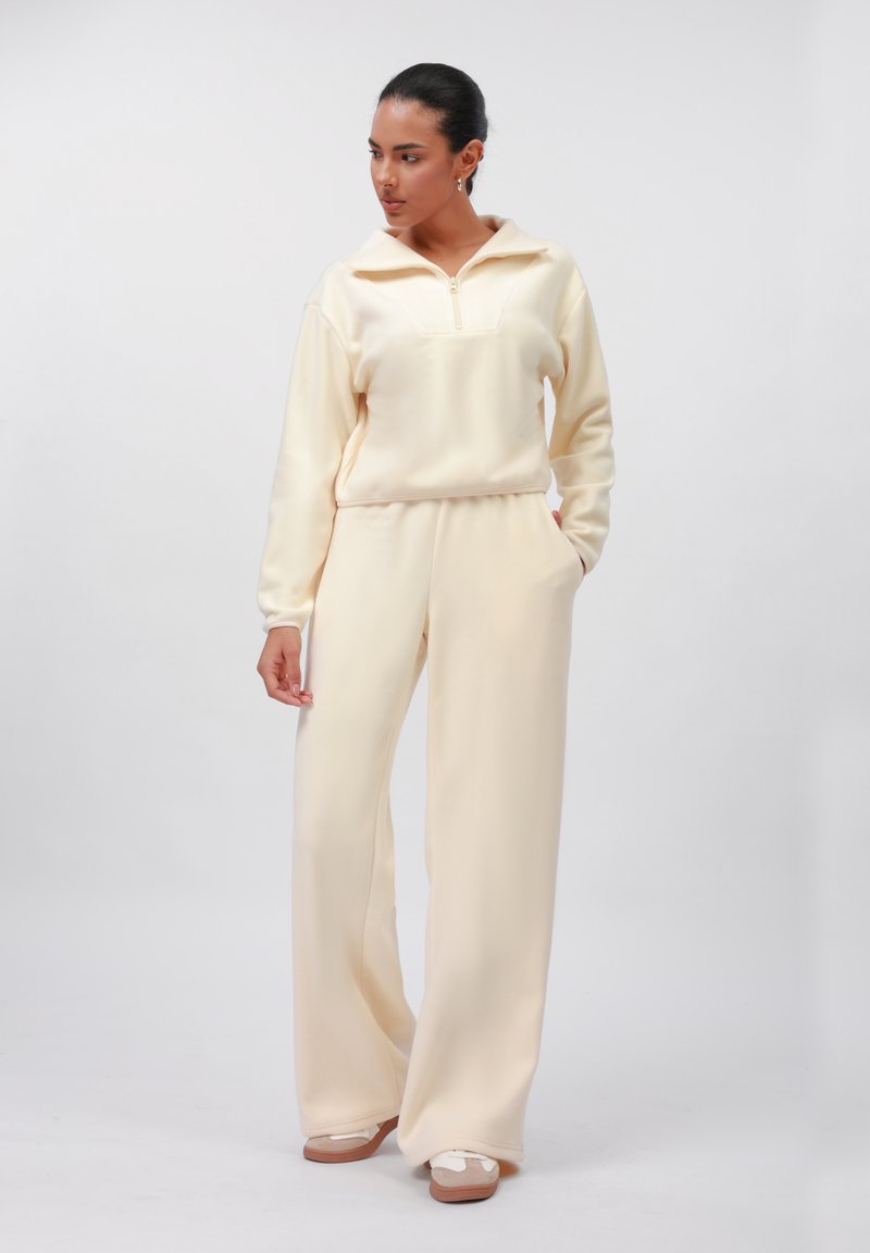 Krem set od loungewear-a napravljen od mekog materijala, s duksericom na zatvarač s ovratnikom i hlačama širokih nogavica, uparen s laganom obućom.