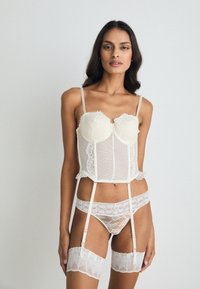 Μποντιού με δαντέλα και πουά σε ελεφαντόδοντο, με ρυθμιζόμενους ιμάντες, ρομαντικές λεπτομέρειες, ζώνες καλτσών και διάφανο ύφασμα. Περιλαμβάνει και καλσόν με δαντέλα που φτάνουν μέχρι το μηρό.