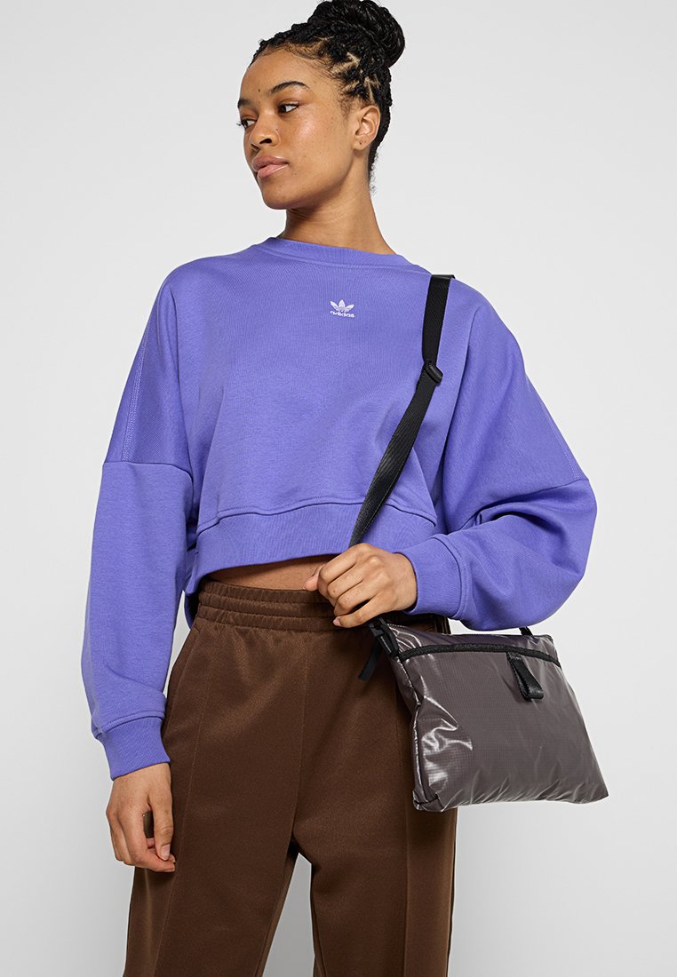 Lila kurz geschnittenes Sweatshirt mit kleinem Logo, kombiniert mit braunen Hosen. Eine silbergraue Umhängetasche mit strukturiertem Design und schwarzen Akzenten.