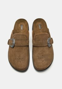 Zoccoli in suede marrone con retro chiuso, adornati con una fibbia decorativa in metallo e un motivo floreale perforato nell'area delle punte.
