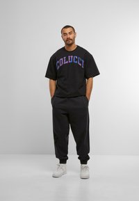 Mand står med hænderne i lommerne iført en sort oversized Colucci t-shirt, sorte joggers og hvide sneakers mod en ensfarvet baggrund.