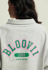 Jasnoszary sweatshirt z dużą grafiką "BLOOVII" w zielonym i różowym kolorze, z mniejszym tekstem "1989" oraz "CAMPUS DREAMER" poniżej.