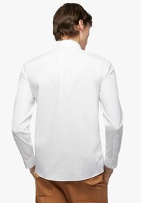 Man in een wit, lange mouw, knoopshirt en bruine broek, staande met handen in zijn zakken, gezien van achteren.