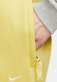 Gula träningsbyxor med en slät yta, sidoficka med dragkedja, och en vit Nike-logga nära fållen. Grå mudd synlig.