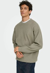 Maglione lavorato a maglia verde chiaro con scollatura a girocollo e polsini a coste. Il tessuto presenta una trama testurizzata. Indossato con jeans blu scuro.