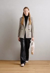 Blazer croisé en motif pied-de-poule, col roulé noir, pantalon large noir et sac à main clair. Fond en bois.