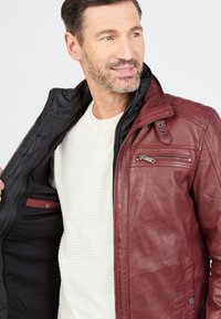 Kastanienbraune Lederjacke mit schwarzer Innenauskleidung, Reißverschlüssen und einem knöpfbaren Kragen, ausgestattet mit Brust- und Seitentaschen.