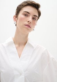 Camicia bianca con colletto a punta, texture morbida e maniche leggermente oversize. Orecchini a bottone argentati aggiungono un dettaglio riflettente.