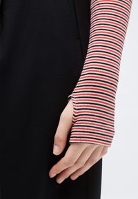 Χέρι με μπλούζα μακρυμάνικη ριγέ σε κόκκινες, λευκές και μαύρες οριζόντιες ρίγες και οπή για τον αντίχειρα, στηρίζεται πάνω σε μαύρο ύφασμα.