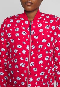 Veste bomber rouge avec un motif floral en blanc et bleu, dotée d'une fermeture éclair à l'avant et de poignets et col côtelés. Texture de tissu lisse.