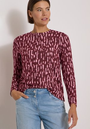 Femme portant un haut à manches longues rose et bordeaux à motifs et un jean taille haute bleu clair, la main dans la poche, regardant sur le côté.