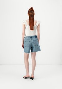 Lichtblauwe denim shorts met een rafelige zoom, gecombineerd met een witte smocked top met ruches aan de mouwen. Het model is van achteren bekeken.