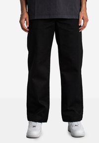 Schwarze weiter Hose aus strapazierfähigem Stoff, mit schlichtem Design und umgeschlagenen Säumen. Getragen mit weißen Sneakers für den Kontrast.