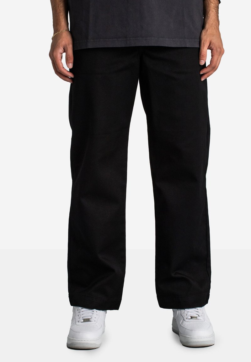 Schwarze weiter Hose aus strapazierfähigem Stoff, mit schlichtem Design und umgeschlagenen Säumen. Getragen mit weißen Sneakers für den Kontrast.