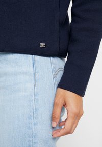 Personne portant un pull bleu marine à manches longues avec un petit logo argenté et un jean bleu clair, main reposant à côté.