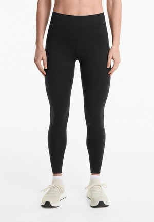 Schwarze, hoch-taillierte Leggings mit glatter Textur. Getragen mit hellen, geschnürten Sportschuhen. Einfaches, figurbetontes Design.