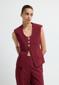 Gilet sans manches bordeaux avec un décolleté V profond, trois boutons dorés et poches avant, fabriqué dans un tissu structuré.