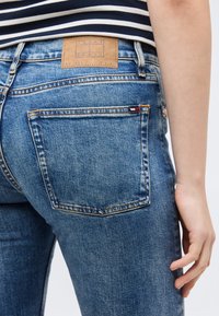 Άτομο που φορά μπλε τζιν από denim με καφέ δερμάτινη ετικέτα μάρκας και μικρή ταμπέλα λογότυπου στην πίσω τσέπη, με το χέρι να ακουμπάει δίπλα στο ισχίο.