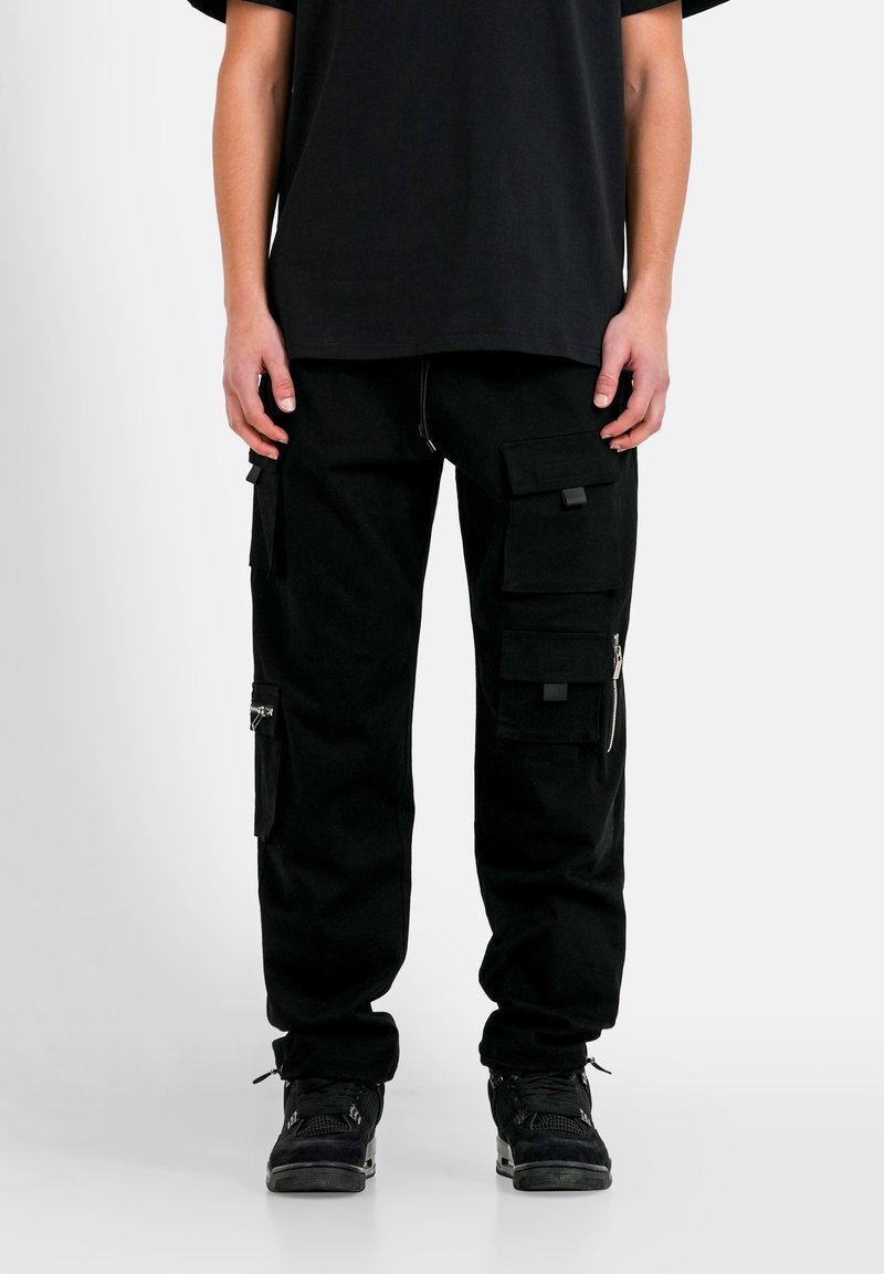Schwarze Cargo-Hosen mit mehreren Taschen, Reißverschlüssen und strukturiertem Stoff. Lockere Passform, schmal zulaufende Bündchen, kombiniert mit einem schwarzen Oversize-T-Shirt und Sneakers.