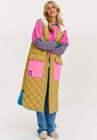 Prešívaná vesta žltej farby s ružovými vreckami a modrými akcentmi. Pod ňou pruhovaná blúzka s dlhým rukávom. Mäkká textúra, dizajn po kolená.