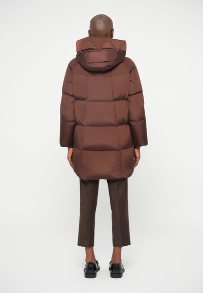 Długa brązowa kurtka puchowa z kapturem, z pikowanymi sekcjami i luźnym krojem, w połączeniu z brązowymi spodniami i czarnymi butami.