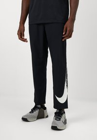 Sorte træningsbukser med et hvidt Nike-logo i penselstrøgsdesign, lavet af letvægtsmateriale, der er glat. Bæres med grå sneakers.