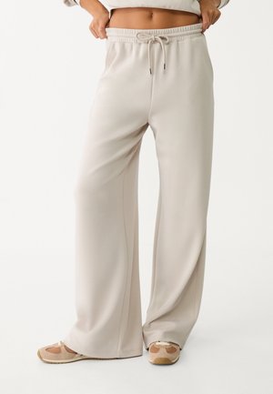 Pantalons larges beiges avec une taille élastique et un cordon de serrage. Fabriqués dans un tissu lisse avec un léger éclat. Complétés par des chaussures sportives.