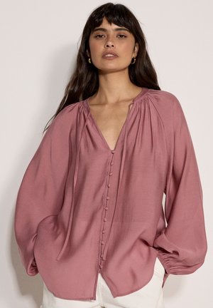 Kobieta o długich ciemnych włosach, nosząca złote kolczyki koła, luźną, pudrowo-różową bluzkę z ozdobnymi guzikami oraz białe spodnie na jednolitym tle.