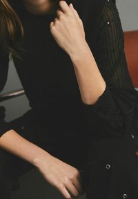 Pull texturé noir avec manches rayées transparentes, silhouette assise reposant la main sur le genou, détails d'accent visibles sur la tenue.