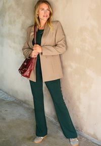 Beige blazer over een donkergroen outfit, met vloeiende broeken. Een rode handtas met studs en grijze schoenen maken de look compleet. Glad muur achtergrond.
