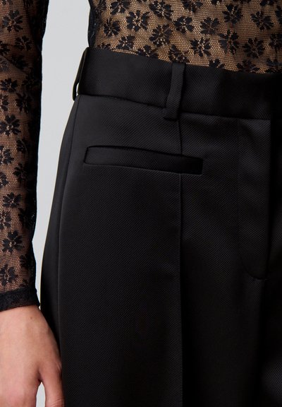 Pantaloni neri su misura con una texture liscia, dotati di una tasca laterale e una vita inclinata, abbinati a un top lungo in pizzo nero trasparente con maniche lunghe.