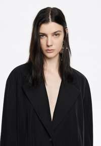 Blazer nero con scollatura a V profonda, realizzato in tessuto liscio. Presenta ampi revers e chiusura a un bottone, con una vestibilità rilassata e una leggera lucentezza.