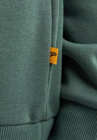 Tessuto felpato verde con texture e dettagli a costine. Presenta un piccolo etichetta gialla del marchio sulla cucitura, a indicare qualità e attenzione ai dettagli.
