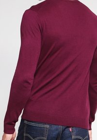 Burgunderfärgad stickad tröja med långa ärmar och ribbad nederkant. Slät textur med en figurnära design, i kombination med mörk denimjeans.