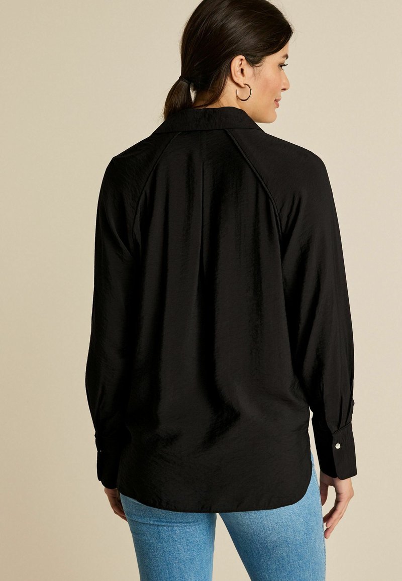 Chemise noire à col avec manches longues, en tissu texturé et poignets boutonnés, portée avec un jean bleu clair, vue de dos.