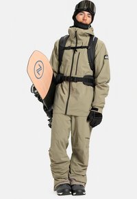 Costum de snowboarding bej, cu o jachetă impermeabilă, pantaloni asortați, mănuși negre și o cască de schi. Ține un snowboard din lemn.