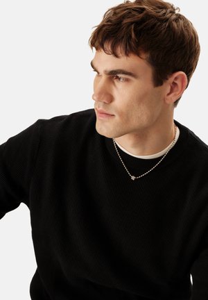 Pull en maille texturée noire avec un col rond, porté par-dessus une chemise blanche à col, agrémenté d'un collier chaîne argenté avec un petit pendentif.