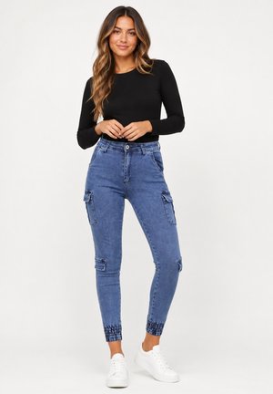 Kvinde iført sort langærmet top, blå cargobukser med elastiske manchetter og hvide sneakers, stående mod en ensfarvet hvid baggrund.