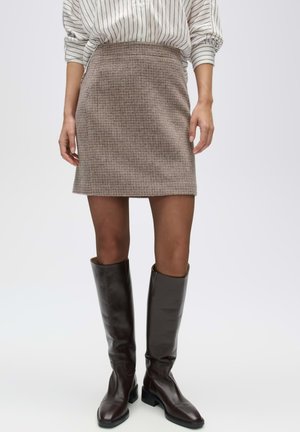 Femme portant une mini-jupe marron à motifs, des collants noirs transparents, des bottes en cuir marron foncé montant jusqu'aux genoux, et un haut blanc à manches longues avec des rayures noires.
