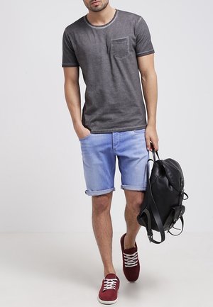 Camiseta de algodón gris con un bolsillo en el pecho, combinada con pantalones cortos de mezclilla azul claro con puños y zapatillas rojas. Mochila negra colgada sobre un hombro.