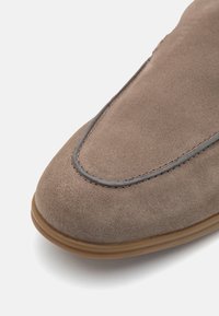 Chaussure en daim brun clair avec un bout arrondi, présentant des coutures grises le long de la couture, et une semelle en caoutchouc beige. Texture lisse, design minimaliste.